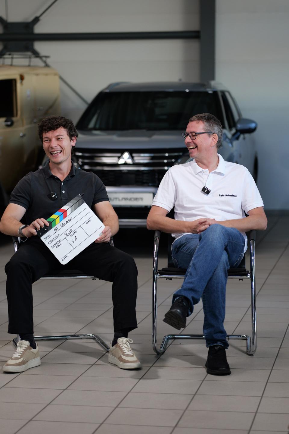Cedric Ruppenthal mit Filmklappe und Benjamin Schneider lachen sich beim Dreh im Autohaus Schneider an